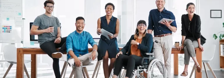 A diverse group of colleagues in an office, smiling and holding devices, with one person in a wheelchair.